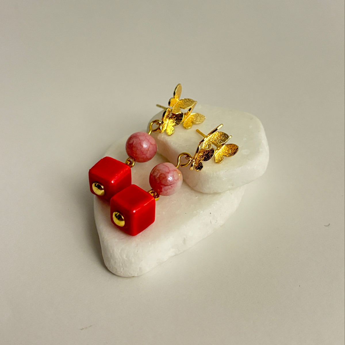 A pair of gold-toned dangle earrings with red cube charms, pink beads, and small gold heart charms, displayed on a white stone stand against a light beige background.