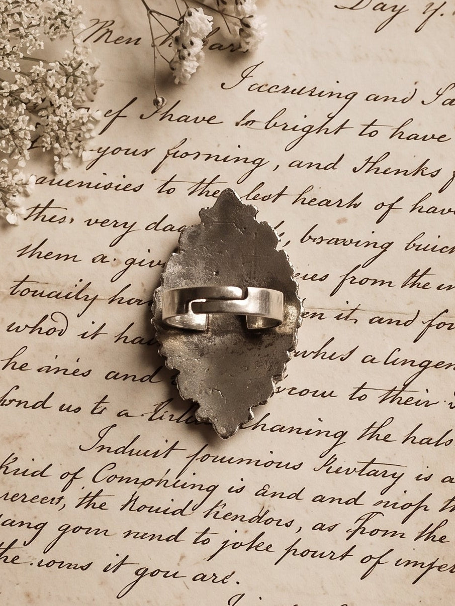 A vintage-style ring with a textured, stone-like center and a silver band, displayed on top of antique handwritten letters with lace and dried flowers on a wooden surface.