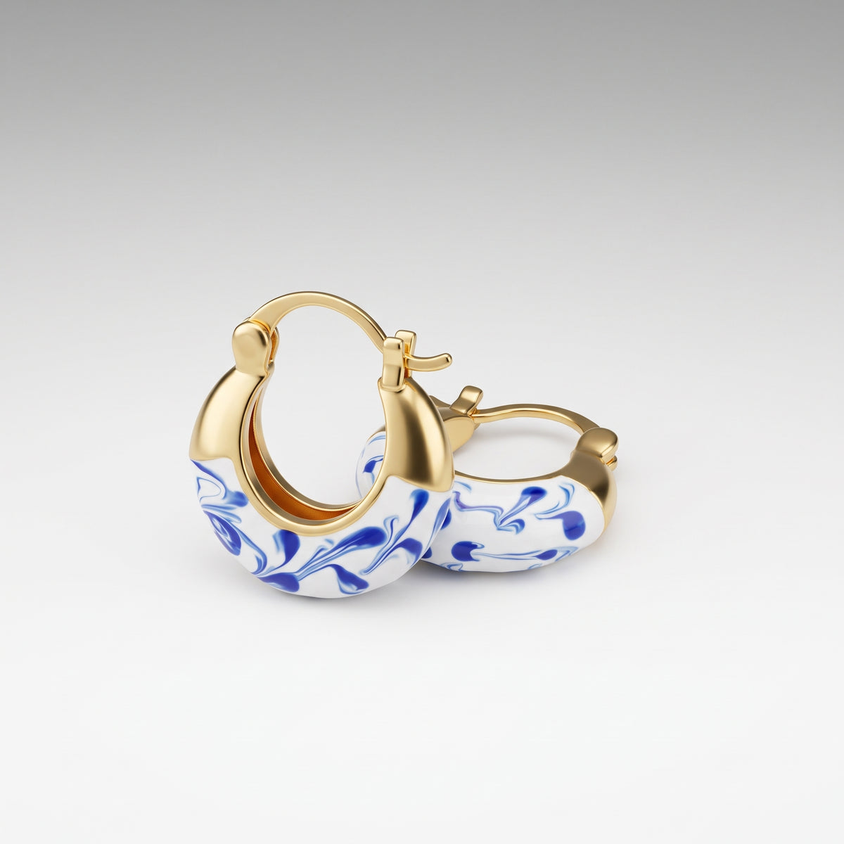 A pair of gold hoop earrings with a white enamel face decorated in a blue floral marbled pattern, shown on a neutral gray gradient background.