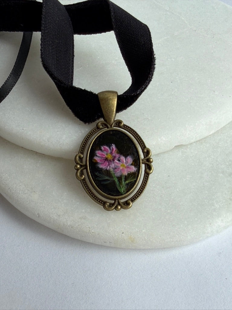 An oval pendant necklace with a black ribbon, featuring a vintage-style bronze frame and a miniature floral painting of pink flowers with green leaves on a black background.