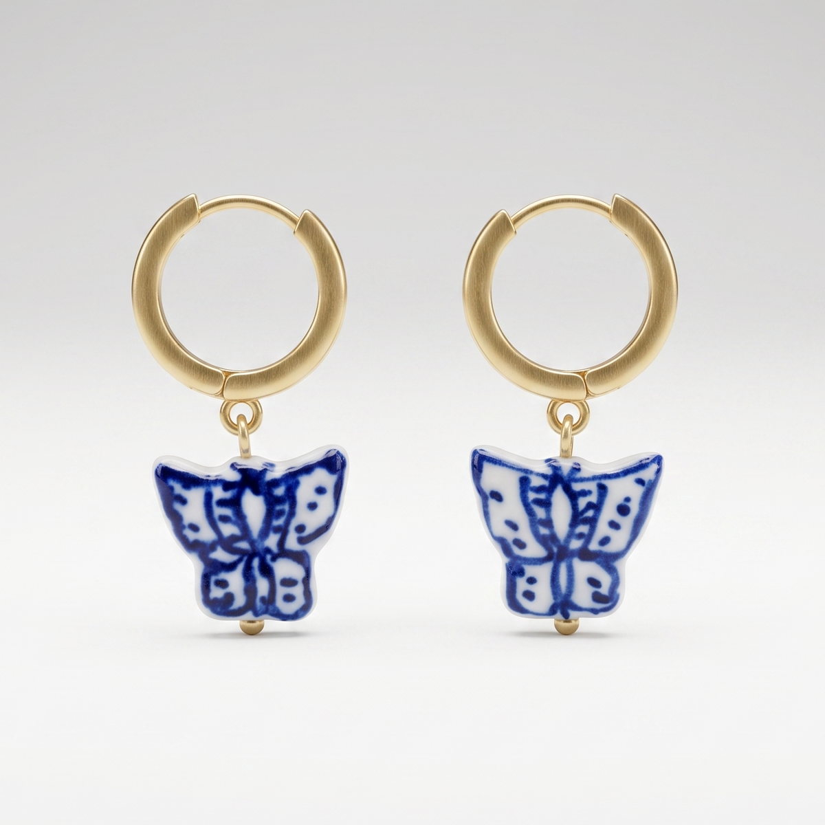 A pair of gold huggie hoop earrings with dangling blue and white butterfly charms, photographed against a seamless light gray studio background.