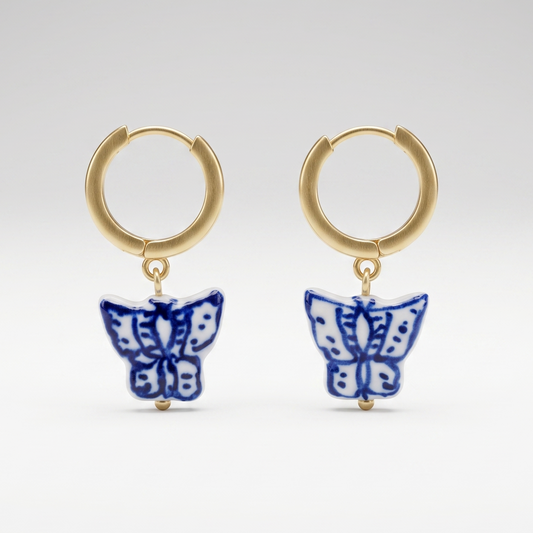 A pair of gold huggie hoop earrings with dangling blue and white butterfly charms, photographed against a seamless light gray studio background.
