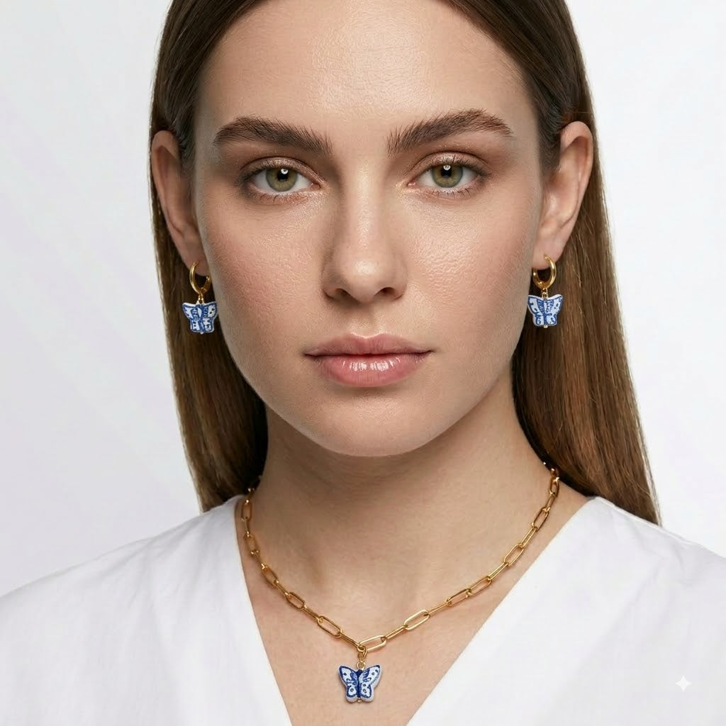 A woman wearing a gold chain necklace with a blue butterfly pendant, matching blue butterfly earrings, and a white top against a light gray background.