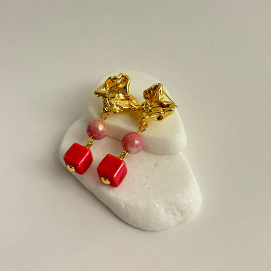 A pair of gold dangle earrings with textured gold tops, pink round beads, and bright red square drop charms, displayed on a small white stone stand against a light gray background.