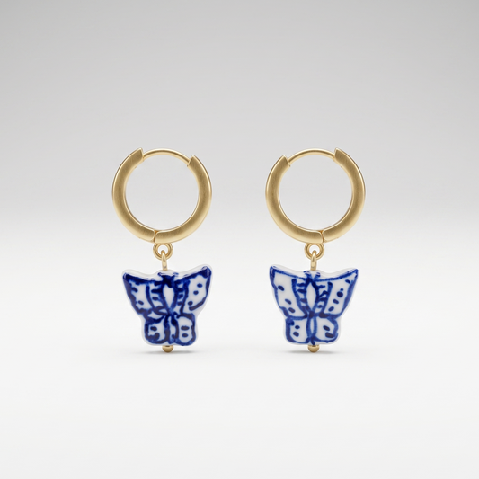 A pair of gold huggie hoop earrings with dangling blue and white butterfly charms, photographed against a seamless light gray studio background.