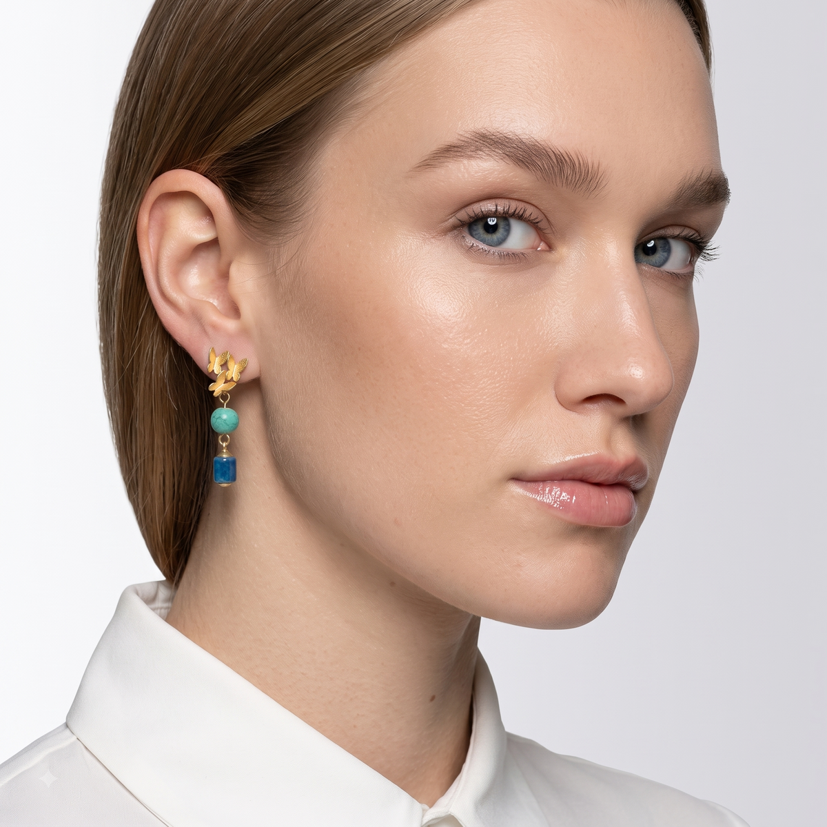 A close-up of a woman wearing a gold dangle earring with a turquoise bead and a blue rectangular charm, against a light gray background.