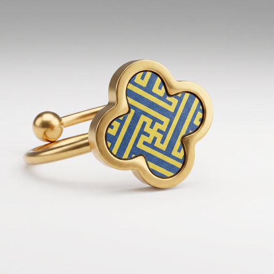 A gold four-leaf clover ring with a blue and yellow geometric pattern inlay, shown on a clean light gray background.