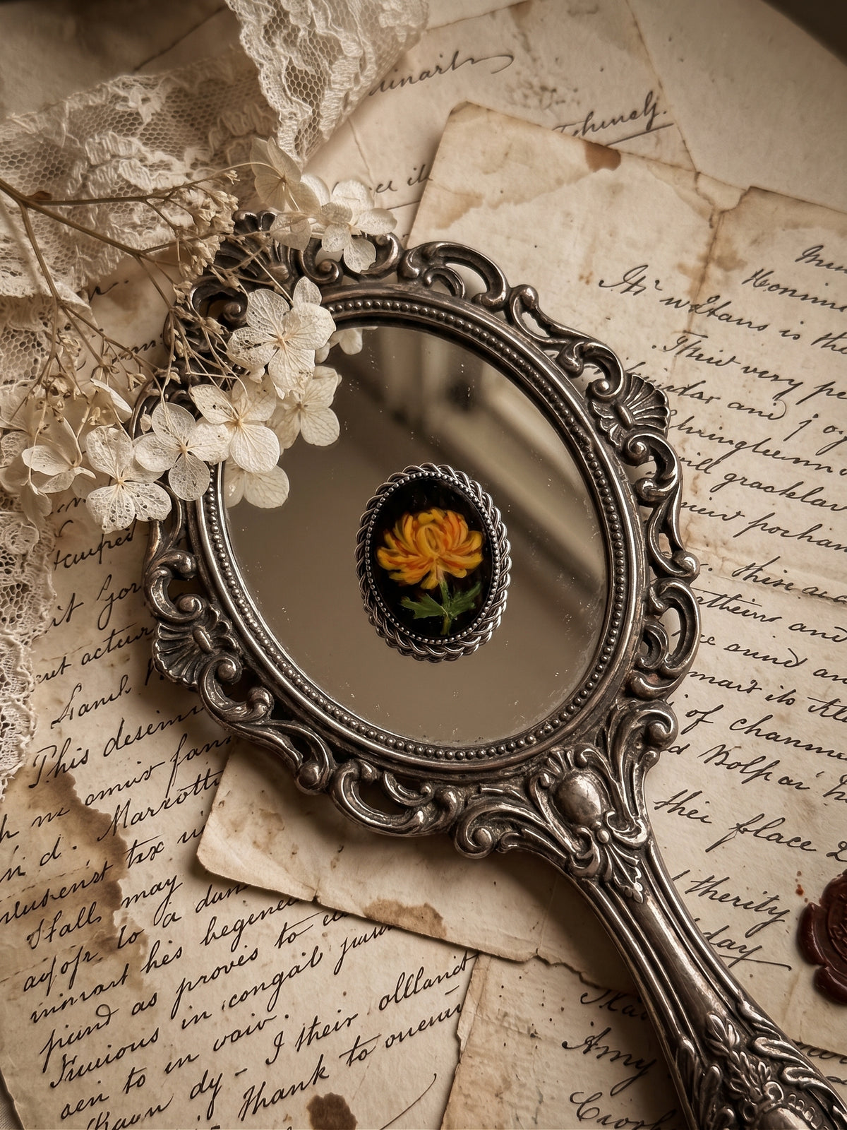 An ornate vintage-style silver hand mirror with a decorative oval frame, reflecting a small yellow flower, styled on aged handwritten paper with lace and dried white flowers.