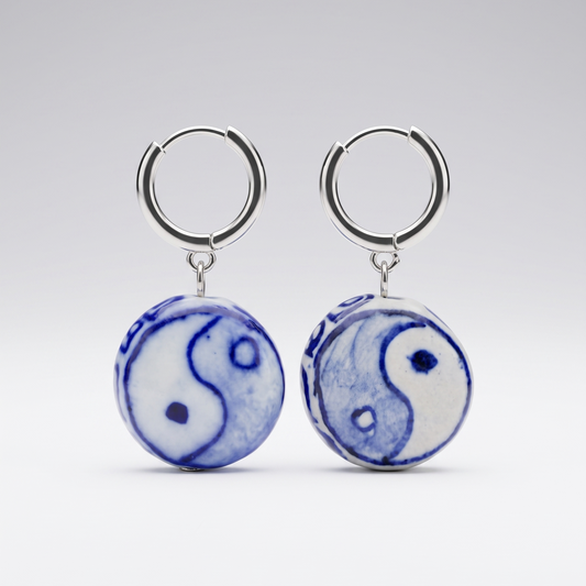 A pair of silver hoop earrings with round ceramic dangles featuring a blue and white yin-yang symbol, shown against a light gray gradient background.