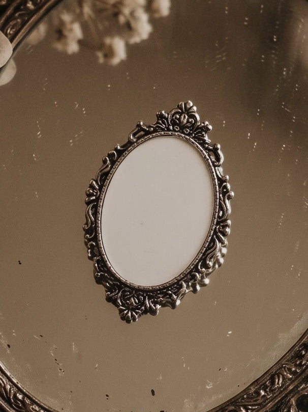 An ornate vintage-style oval hand mirror with a heavily carved antique silver frame, styled on top of aged handwritten letters, lace, and dried flowers.