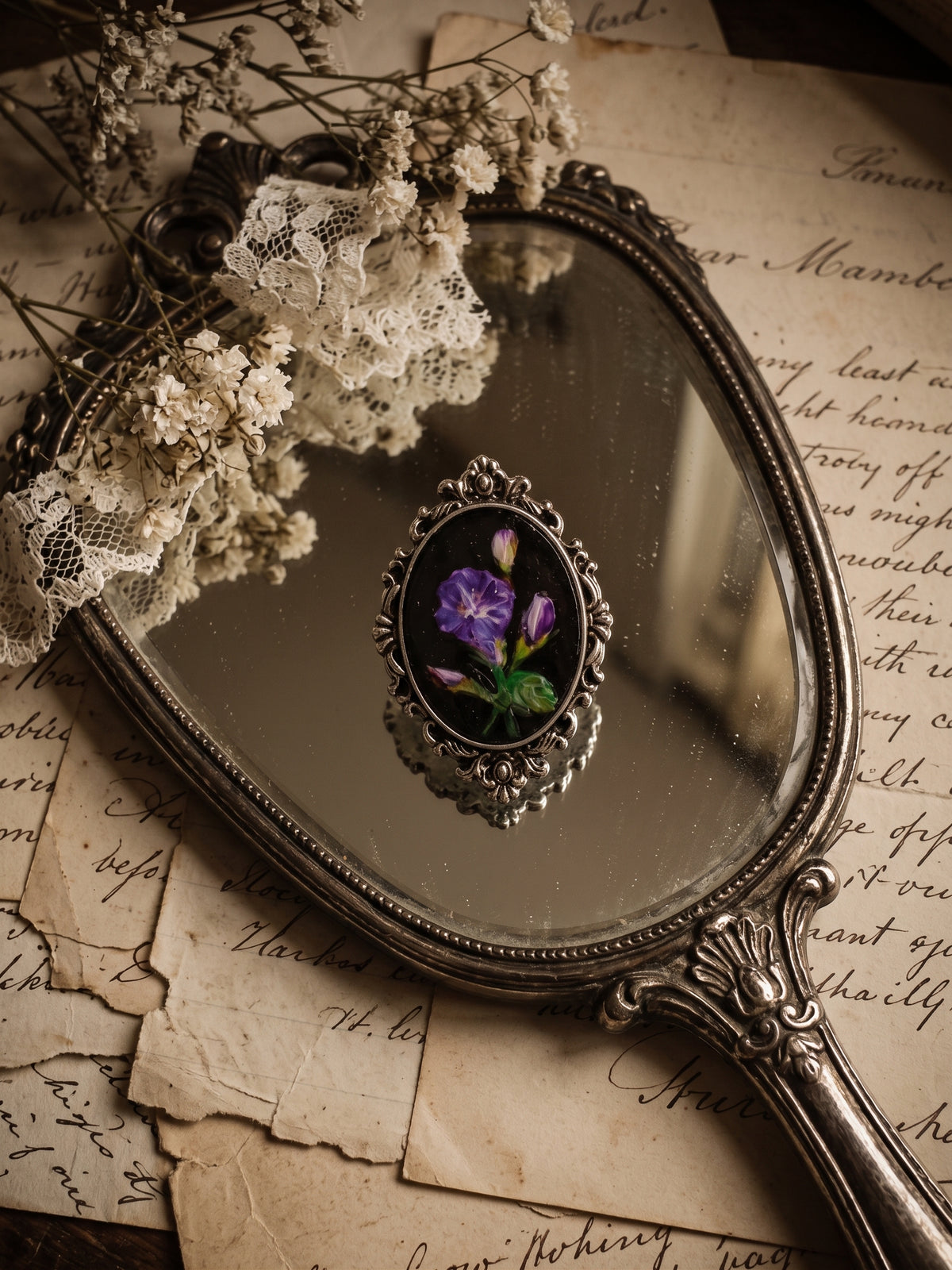 An ornate vintage-style silver mirror with a floral purple and green cabochon in the center, styled on aged handwritten paper with lace and dried flowers.