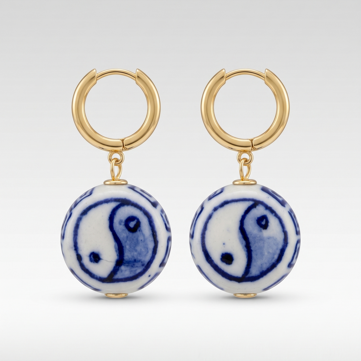 A pair of gold hoop drop earrings featuring round white ceramic pendants with a blue and white yin-yang symbol, set against a light gray gradient background.