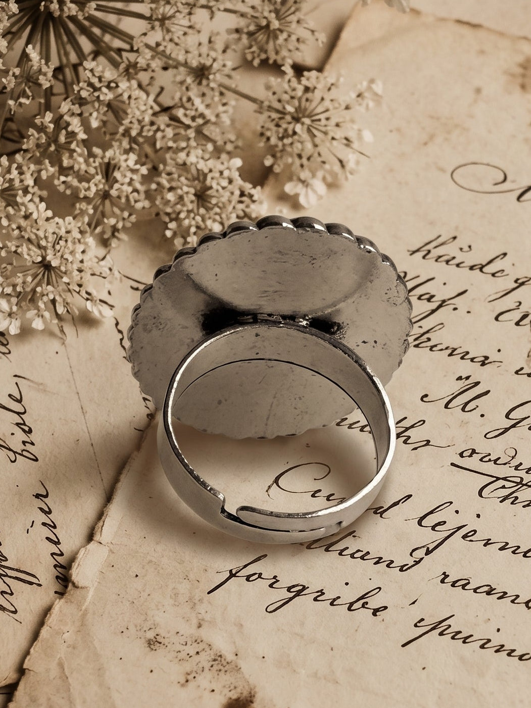 A silver-toned metal ring with a textured, hammered circular face and a smooth band, displayed on top of vintage, handwritten letters and dried white flowers.