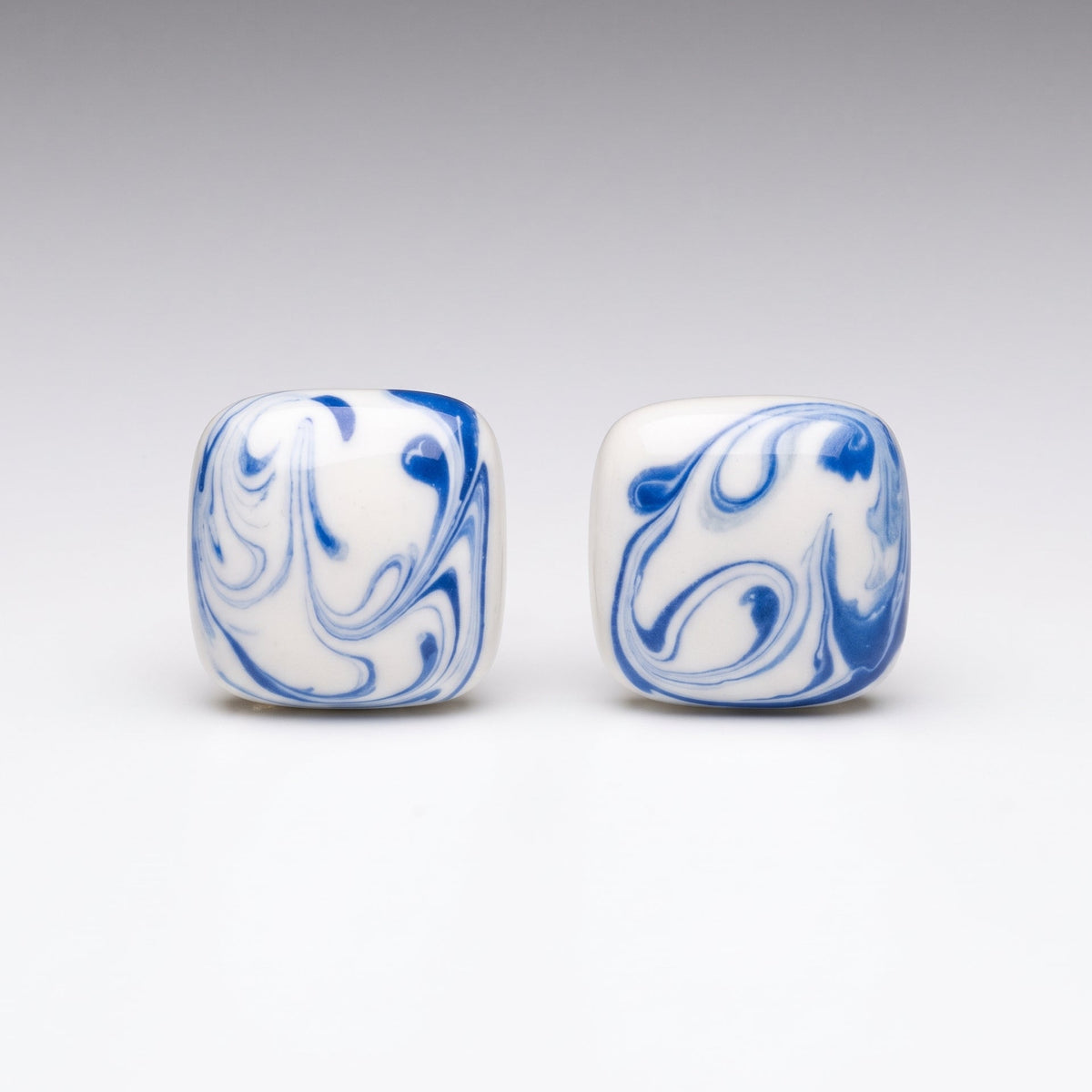 Two square white beads with blue marbled swirl patterns, displayed side-by-side on a light gray gradient background.