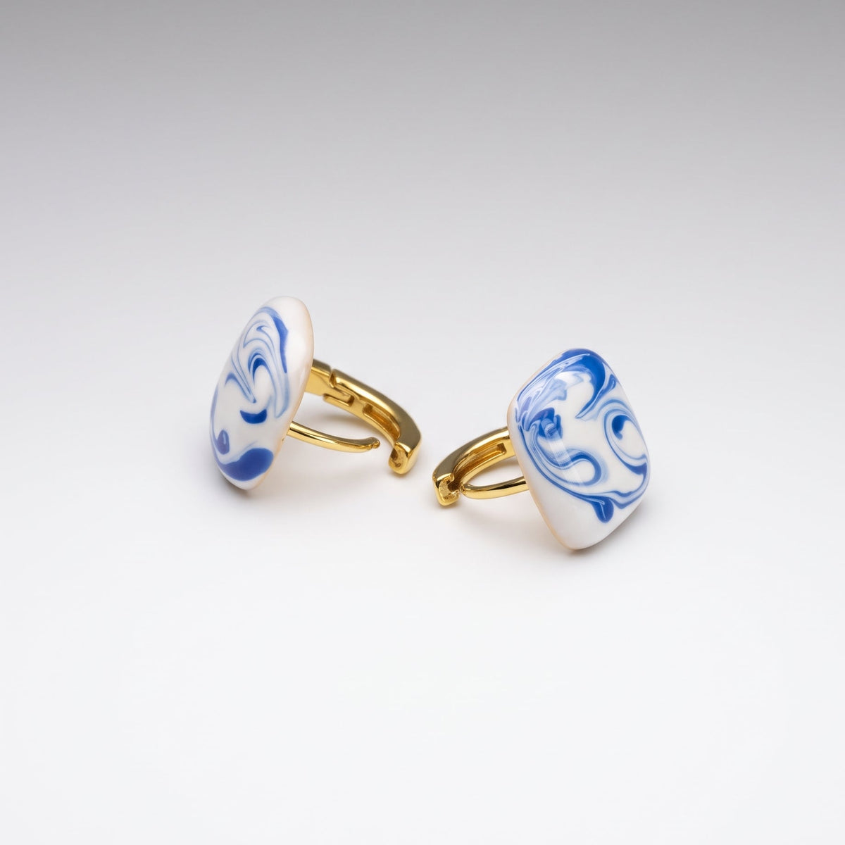A pair of men's cufflinks with oval white enamel faces featuring blue floral scrollwork, attached to gold-tone metal bars, set against a clean white to light gray gradient background.