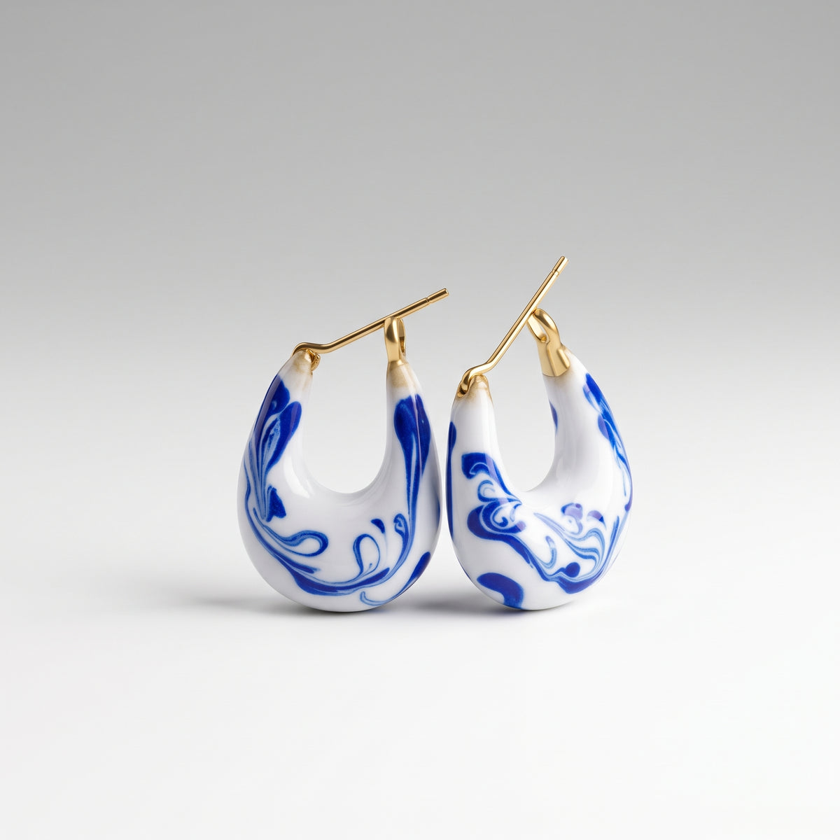 A pair of gold hoop earrings with white teardrop-shaped drops decorated in a blue marbled pattern, shown against a neutral gray gradient background.