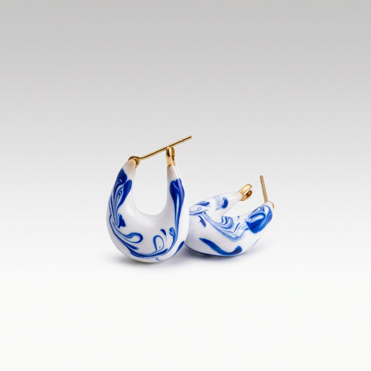 A pair of crescent-shaped earrings with a white base, blue marbled swirl pattern, and gold-tone posts, shown on a clean light gray gradient background.