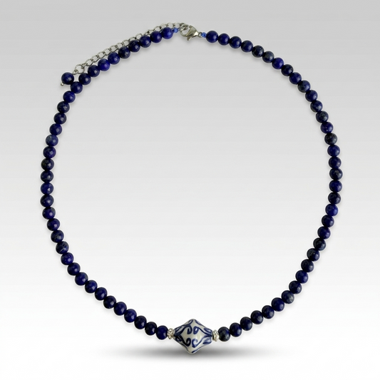 A beaded necklace featuring deep navy blue beads with a central diamond-shaped glass pendant decorated with blue and white floral patterns, set against a light gray gradient background.