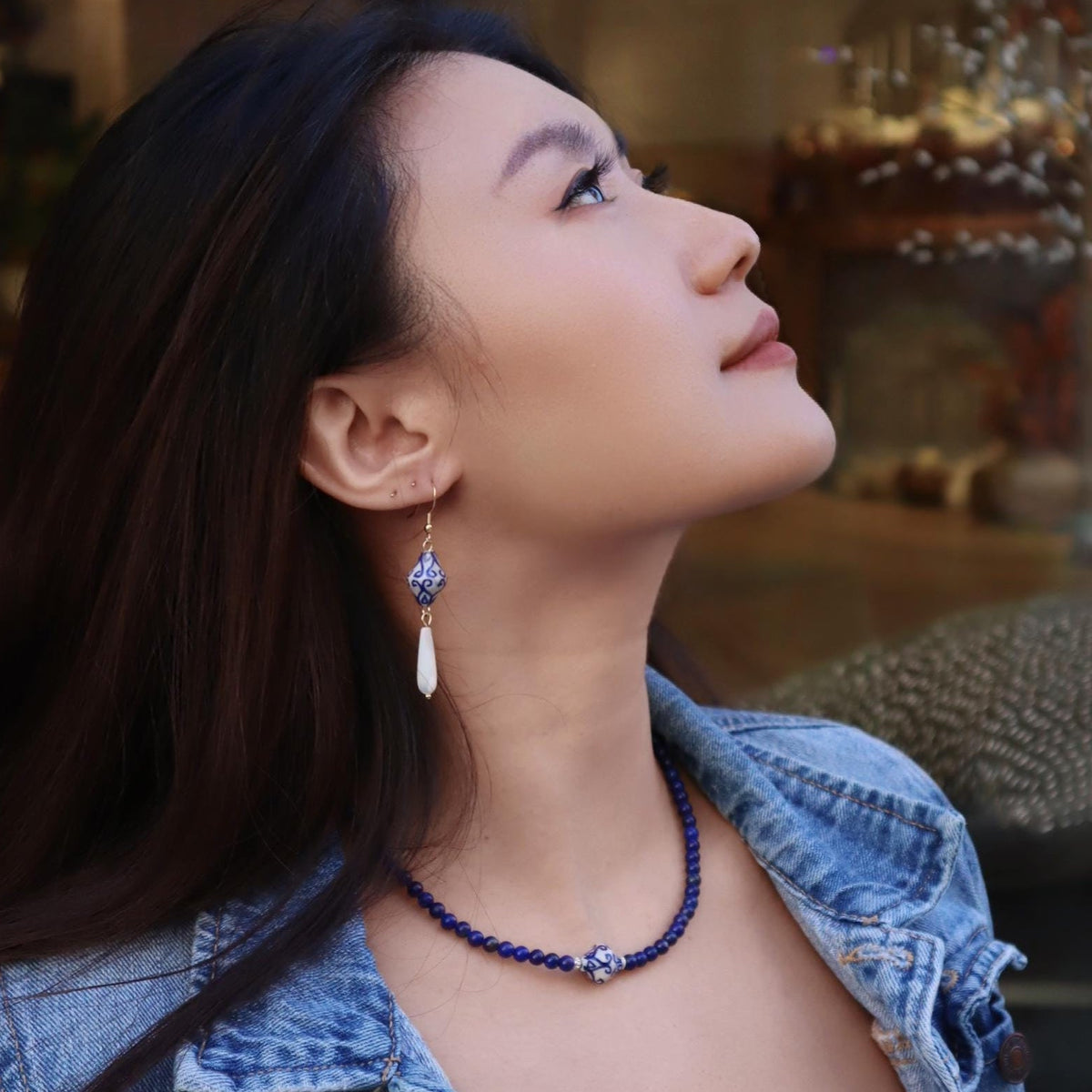 Woman wearing blue earrings and a blue necklace, and denim jacket looking up outdoors.