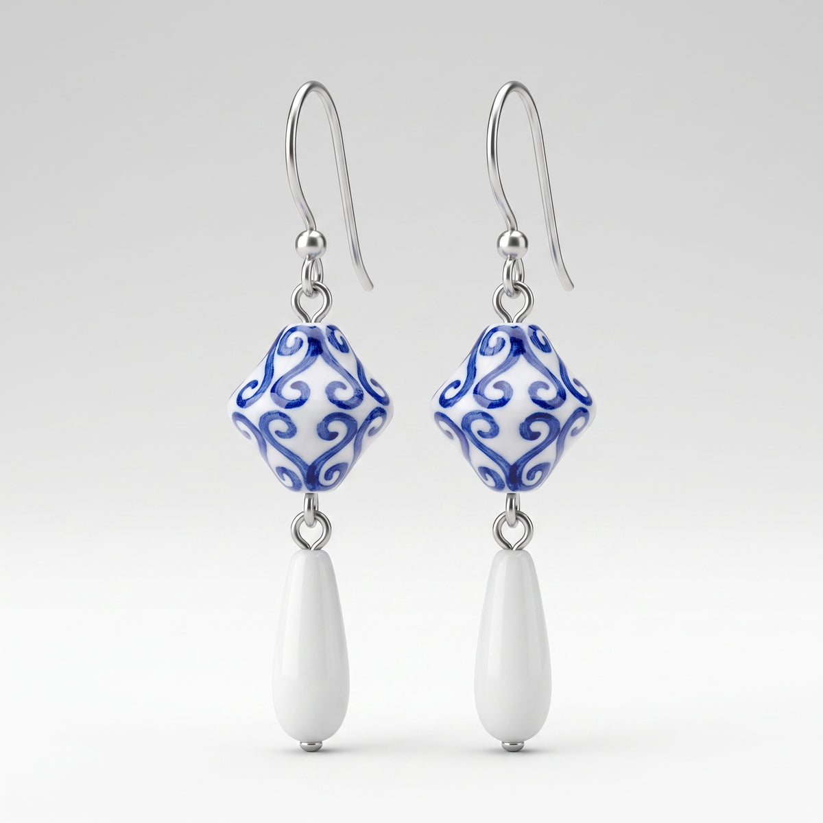 A pair of drop earrings featuring a blue and white patterned ceramic bead with a silver fishhook ear wire and a dangling white teardrop bead, shown against a light gray gradient background.