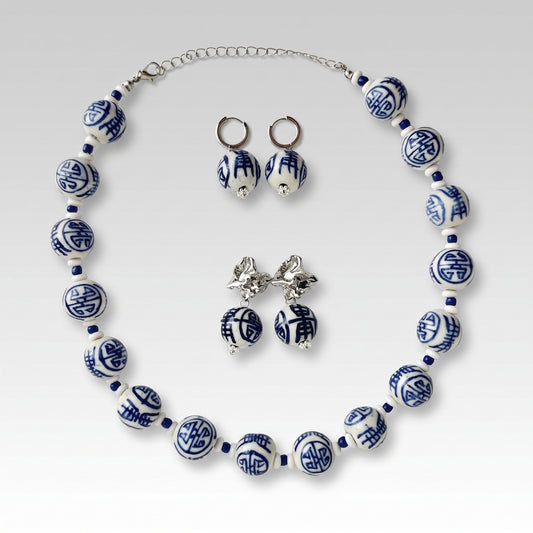 A jewelry set featuring a beaded necklace, matching hoop earrings, and stud earrings, all styled in a classic Chinese porcelain pattern of blue and white with silver metal findings, displayed on a light gray gradient background.