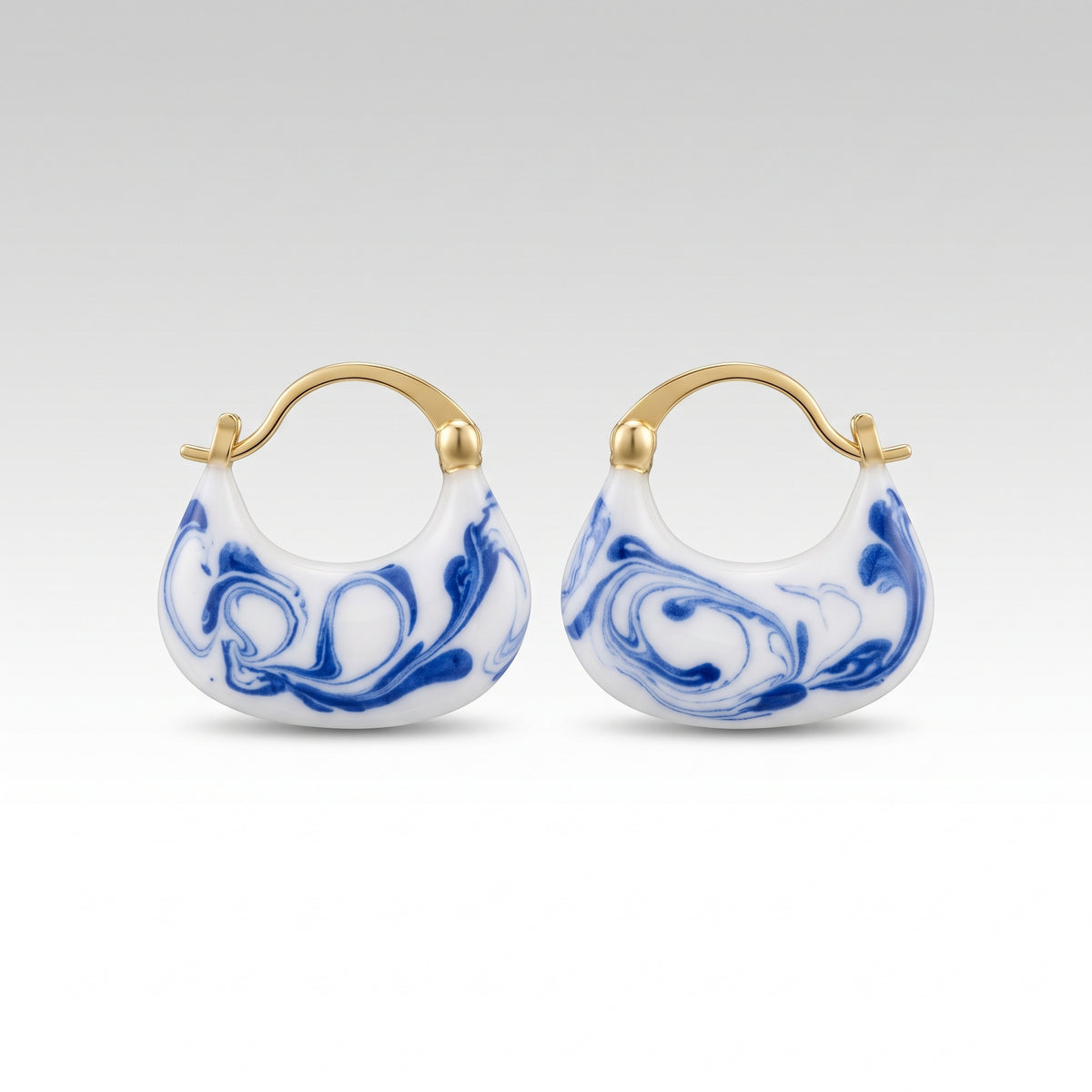 A pair of small crescent-shaped hoop earrings with gold-tone metal tops and blue-and-white marbled enamel, shown on a clean light gray background.