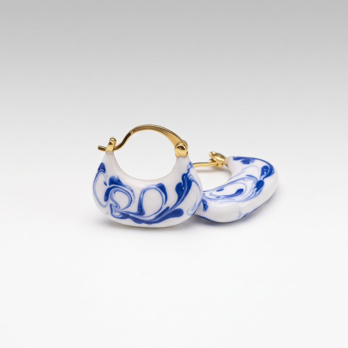 A pair of small hoop earrings with a white base, blue marbled swirl pattern, and gold-tone metal hoops, shown on a clean light gray gradient background.