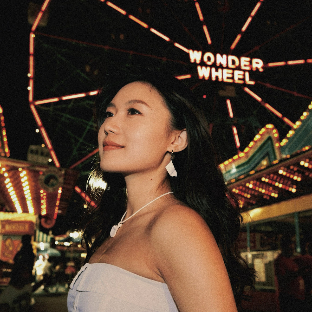 A woman in a light lavender strapless dress stands in front of a brightly lit Ferris wheel at night