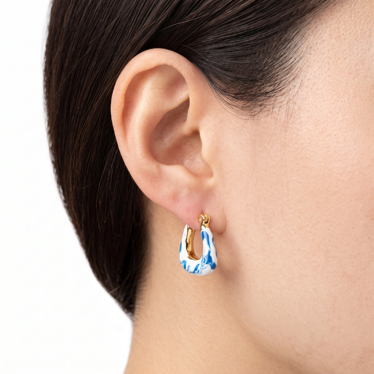 A close-up of a person's ear wearing a small hoop earring with a blue and white marbled pattern and gold metal trim.