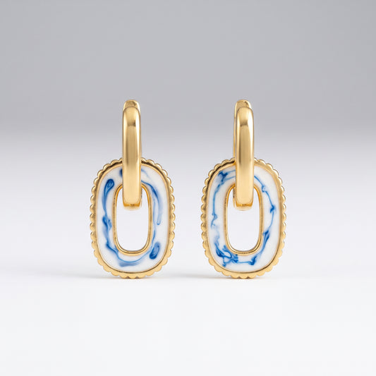 A pair of gold-toned oval drop earrings with a white enamel center featuring blue marbled patterns, set against a clean light gray background.