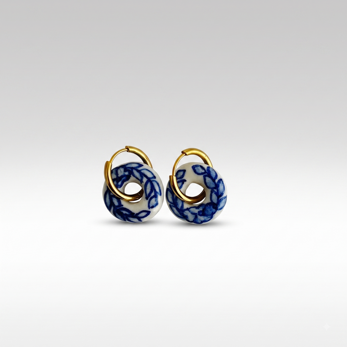 A pair of gold hoop earrings with circular white enamel inserts decorated in a blue floral pattern, shown against a clean light gray gradient background.