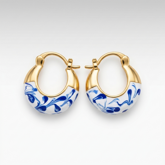 A pair of gold hoop earrings with white enamel inlays featuring a blue floral marbled pattern, shown on a light gray gradient background.