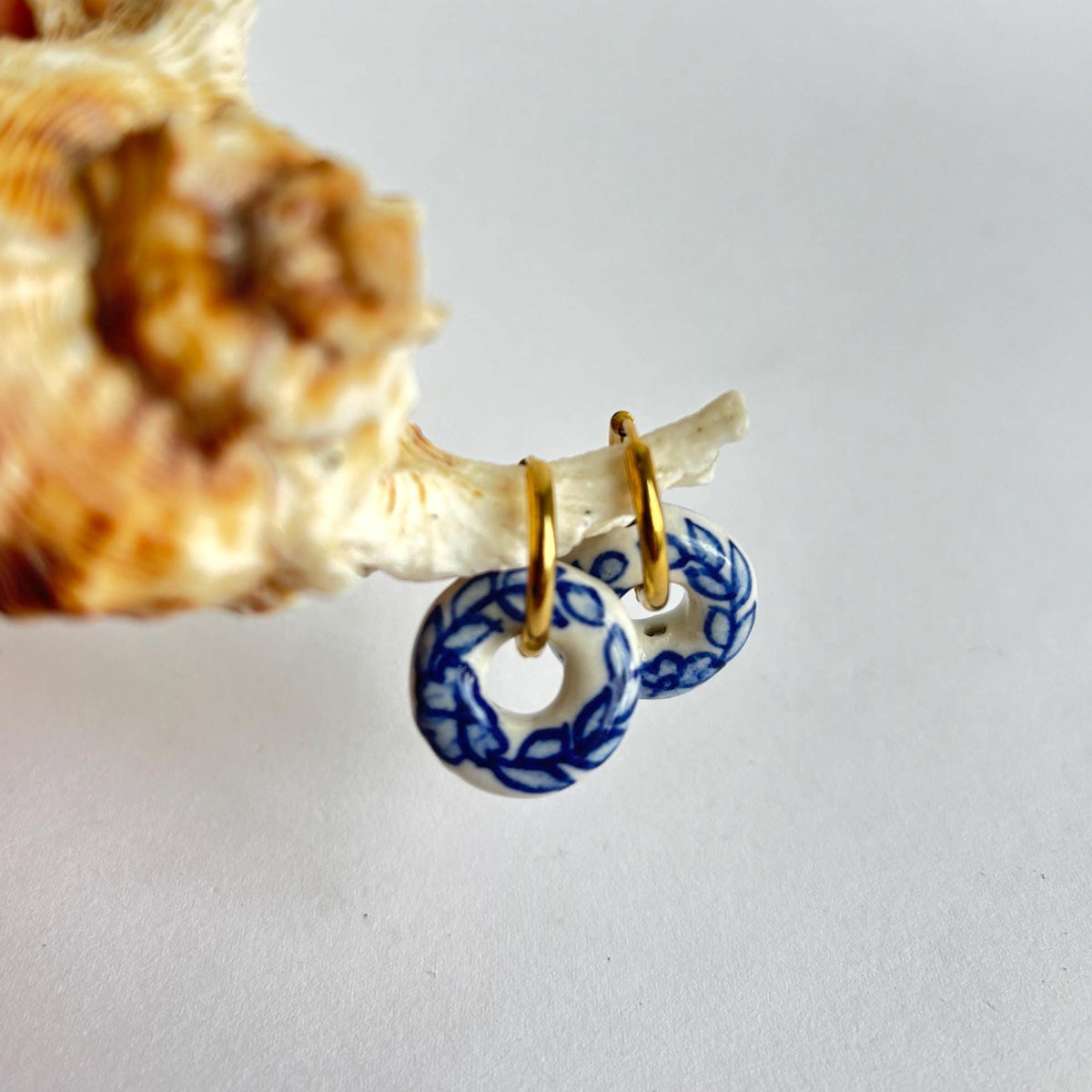 A pair of small hoop earrings with blue and white patterned circular charms, held against a white background next to a seashell.