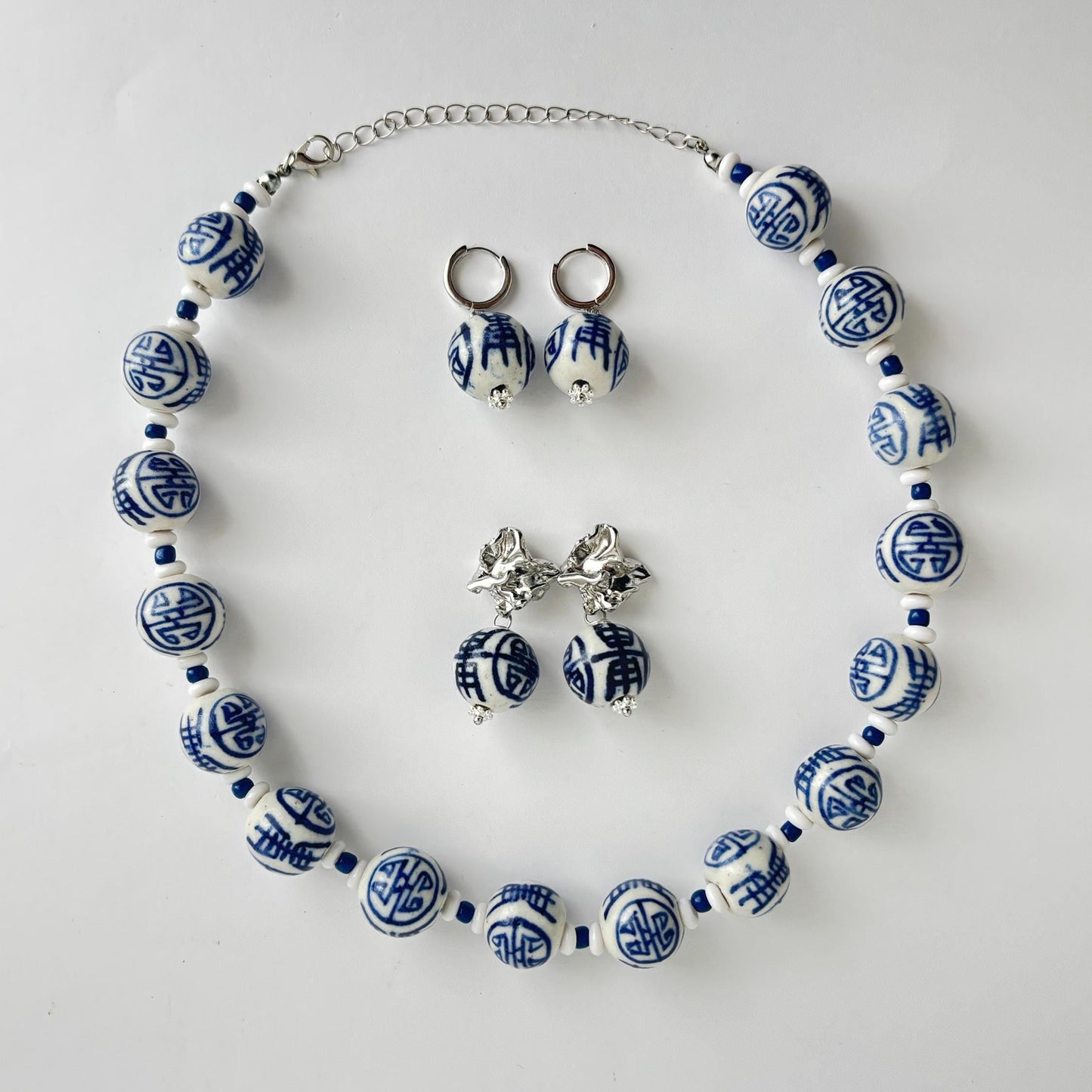 A jewelry set featuring a beaded necklace, matching drop earrings, and stud earrings with blue and white patterned ceramic beads and silver-tone metal findings, arranged on a light gray background.