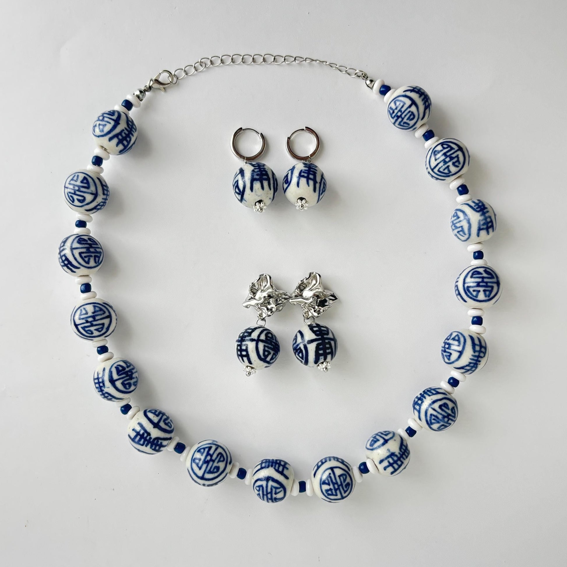 A jewelry set featuring a beaded necklace, matching drop earrings, and stud earrings with blue and white patterned ceramic beads and silver-tone metal findings, arranged on a light gray background.