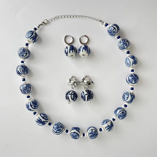 A jewelry set featuring a beaded necklace, matching drop earrings, and stud earrings with blue and white patterned ceramic beads and silver-tone metal findings, arranged on a light gray background.