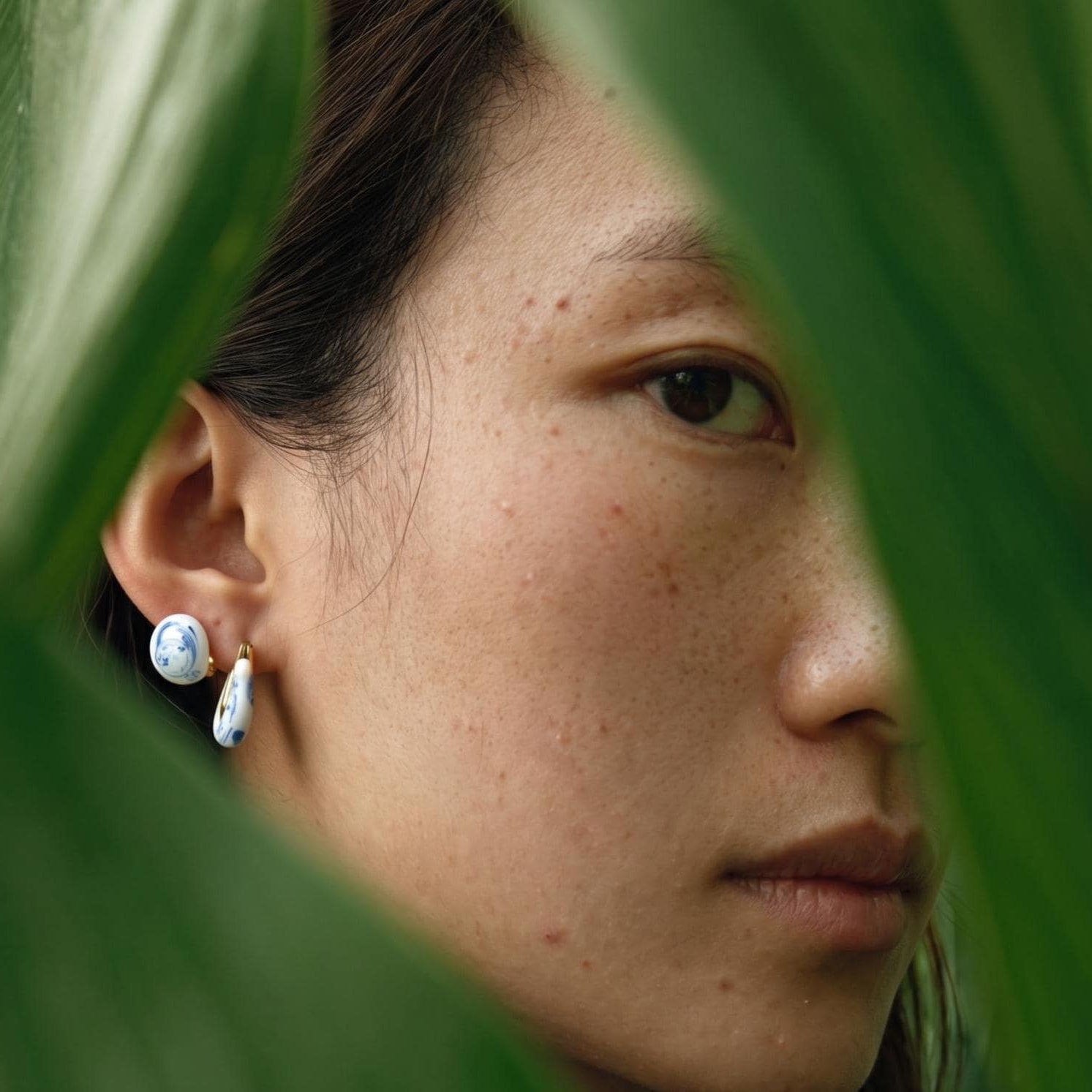 Close-up of a person wearing blue marble stud earrings with green leaves in the foreground