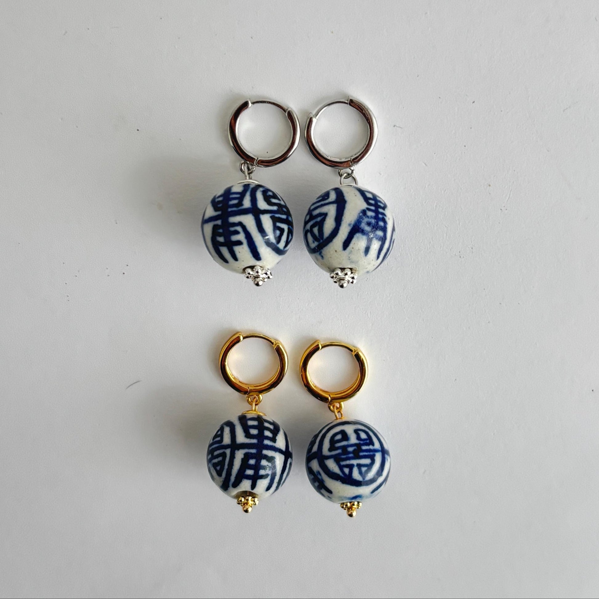 Two pairs of drop hoop earrings displayed on a plain light background. The top pair has silver-toned hoops with white ceramic beads featuring blue Chinese characters and tiny clear rhinestones.