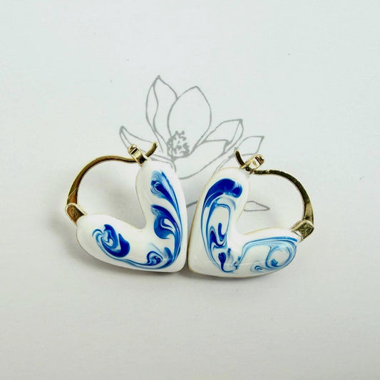 A pair of heart-shaped earrings with white enamel, blue marbled swirl patterns, and gold-tone hoops, displayed on a white background with text reading 'Elegance and Grace, Nature's Embrace'.