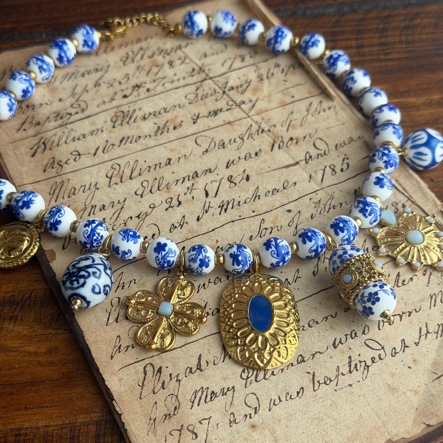 A vintage-style beaded necklace with blue and white patterned beads, gold-tone filigree charms, and a central blue stone pendant, displayed on top of an old handwritten paper document over a wooden surface.