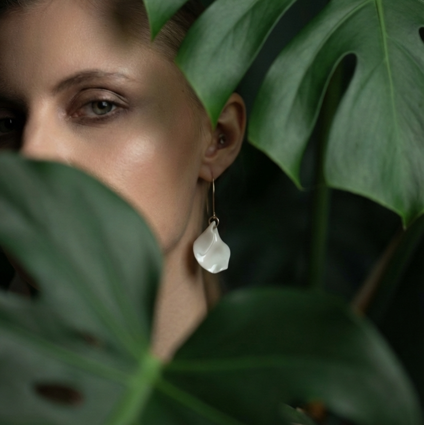 A close-up lifestyle shot of a woman wearing a delicate white teardrop dangle earring, framed by large green monstera leaves.