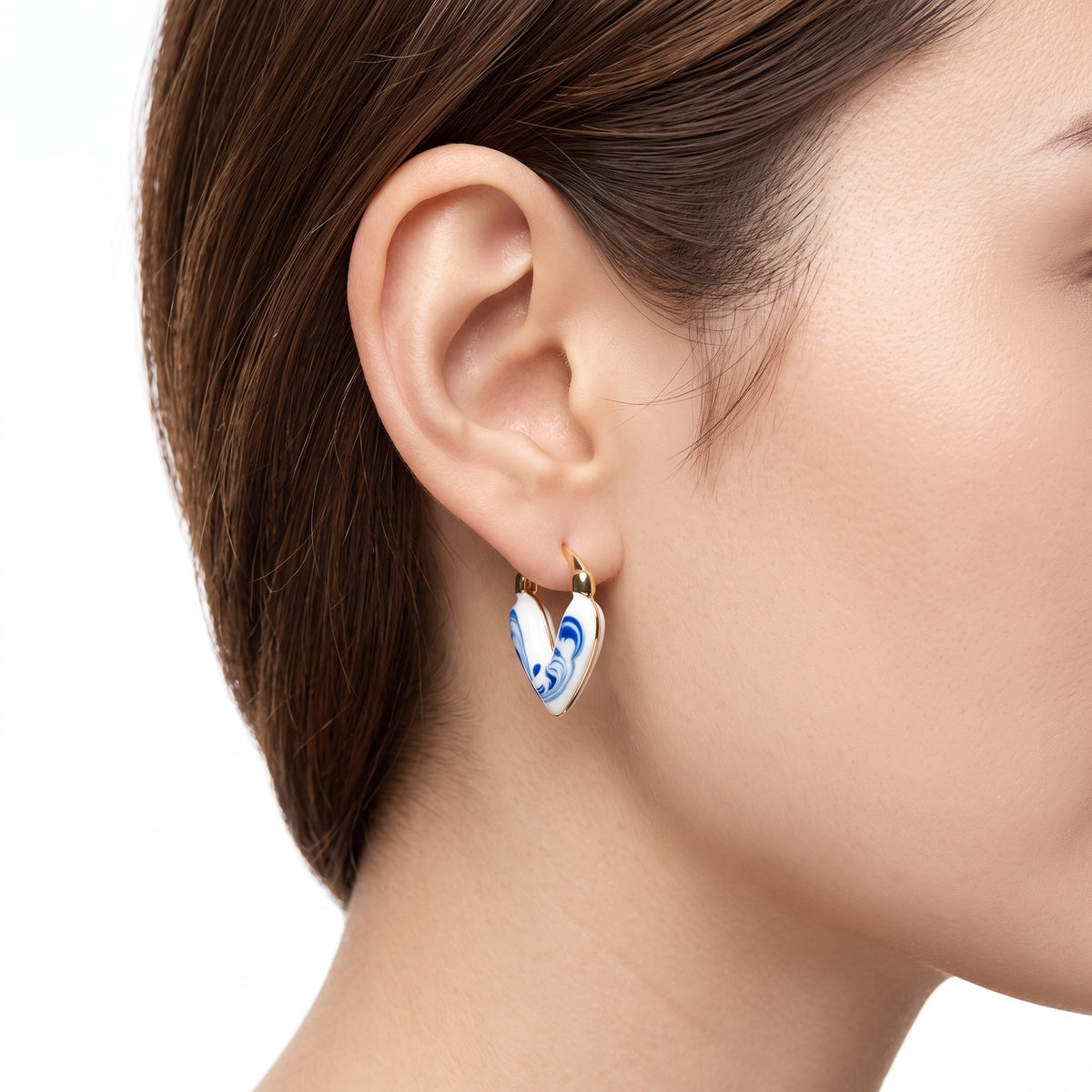 A close-up of a woman's ear wearing a small gold hoop earring with a white heart-shaped drop featuring blue marbled patterns.