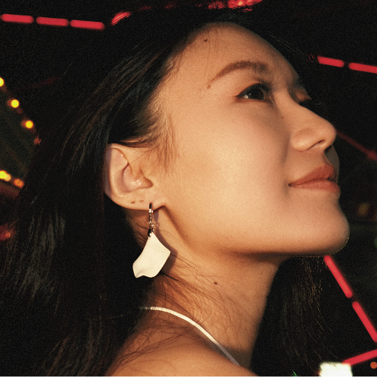 A woman in a white strap top wearing a white petal dangle earring, photographed in a warm-toned night scene with a red illuminated Ferris wheel in the background.