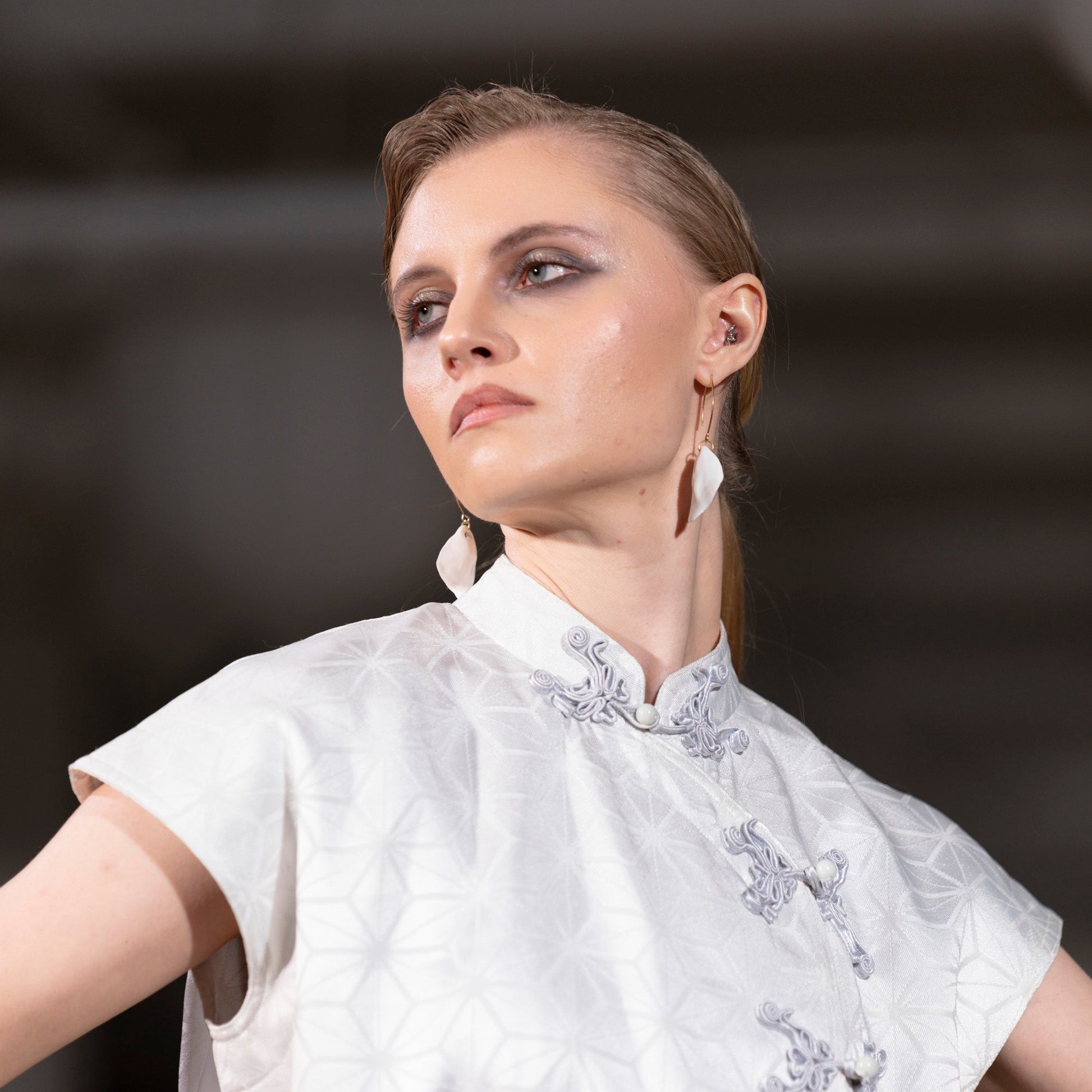 A model wearing a white short-sleeved dress with a high collar, subtle geometric texture, and silver-gray embroidered details on the front, photographed against a dark, blurred background.
